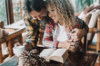 © simona - Romantic indoor scene of happy adult couple, woman with curly hair sitting at table reading book while man with beard embraces her from behind, both smiling and enjoying affectionate morning together