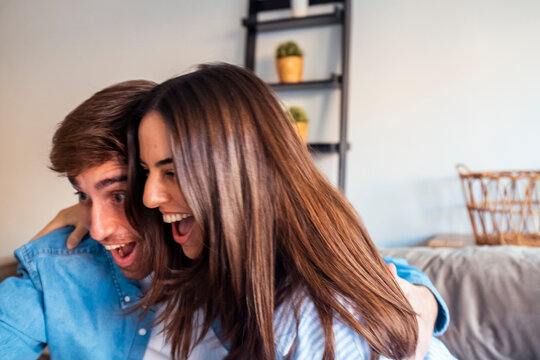 A young couple sitting on the sofa in their living room, watching a series together in the afternoon, enjoying on-demand content and streaming entertainment while spending quality time at home