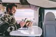 © simona - An adult man sitting inside a modern motorhome by the beach, working remotely on his laptop and using his phone for hotspot and data roaming to stay connected and complete work tasks while traveling