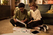 © pressmaster - Two people sitting on floor, closely examining financial documents and discussing planning. Papers, forms, tablets scattered around indicating collaborative effort in budgeting