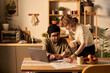 © pressmaster - Young couple working on a project together at home using laptop and documents on a kitchen table. Seminal light illuminating modern kitchen with various house plants is visible