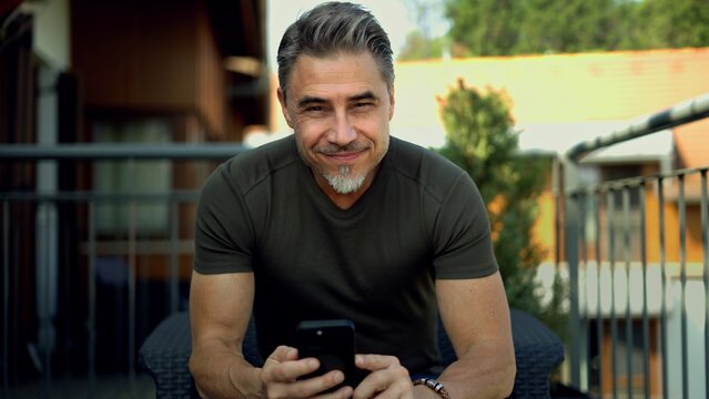 Fototapete -  - Mid adult man working with phone, sitting on balcony or terrace outdoor, checking e-mail, reading news feed, posting on social media. Relaxed, confident happy smile. Businessman in home office.