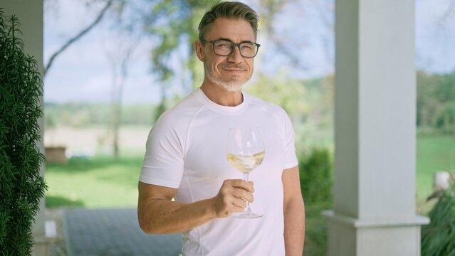 Fototapete -  - Portrait of happy mid adult man drinking white wine on country house terrace. Winemaker standing on porch of winery tasting from glass, smiling. Confident satisfied healthy male in 50s. Lifestyle.