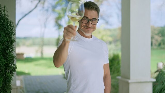 Fototapete -  - Happy man raising a toast with white wine on house terrace, enjoying country life. Winemaker celebrating success of harvest, cheering with glass. Portrait of smiling middle aged man greeting guest.