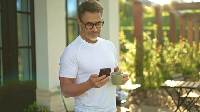 Fototapete -  - Relaxed mature man standing on terrace in garden drinking coffee checking smartphone in morning sun. Businessman outdoor at home, perfect start of a long business day. Life of successful entrepreneur.
