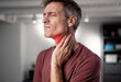 © Nedrofly - A man holds his neck with a pained expression, indicating discomfort in his throat area. The background displays a contemporary living space, suggesting a casual environment
