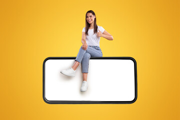 Aufkleber - Positive casual millennial lady sitting on big horizontal smartphone with blank screen and showing thumbs up ok gesture, recommending newest mobile application, isolated on orange studio background
