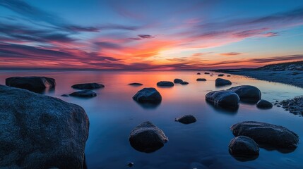Poster - A breathtaking twilight sky reflecting vibrant colors over calm water, with large rocks scattered along the peaceful shoreline, creating a serene atmosphere.