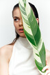 © VICHIZH - Young woman concealing her face behind a green tropical leaf, showcasing elegance and serenity in a minimalist setting with a focus on nature.