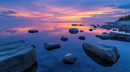 Poster - A stunning twilight scene where the sky colors are mirrored on the calm water, with rocks scattered along the shoreline creating a tranquil, scenic view.