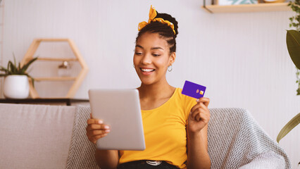 Poster - E-Commerce Concept. Smiling African American Woman Shopping Online Using Digital Tablet And Showing Credit Card, Sitting On The Couch In Living Room At Home. Internet Shop Offers And Sales