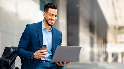 Wall Mural - Positive arabic guy in suit project manager sitting next to business center, drinking coffee and using laptop, getting ready for business meeting or startup presentation, panorama with copy space