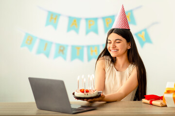 Indian teenage girl in festive hat having birthday party online, sitting near laptop with gift boxes and cake. Happy adolescent celebrating holiday on web, chatting with friends or family on internet