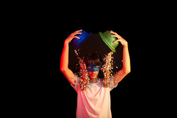 Young man in white shirt wearing carnival mask having fun with two hats and paper confetti. Isolated on black background.