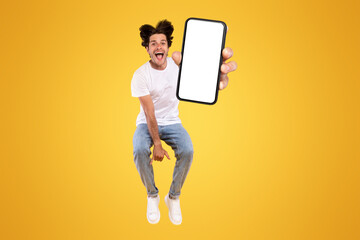 Poster - Handsome bearded young man recommending new mobile application, vertical collage. Excited guy holding smartphone, showing white blank screen, jumping up over orange yellow studio background