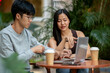 © bongkarn - An attractive Asian woman discussing work at an outdoor table in a coffee shop, working remotely.