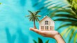 © muhamad - Dream Home Concept. Hand Holds Miniature House And Palm Tree On Blue Background. Vacation Property And Real Estate Investment.
