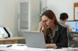 © crizzystudio - Young businesswoman feeling eyestrain, rubbing her nose bridge while working on laptop computer at her desk in modern office, suffering from headache or blurry vision after long computer work