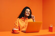 © Jacob Lund - Gen z woman in an orange monochromatic setting using a laptop and holding drink, smiling and looking up