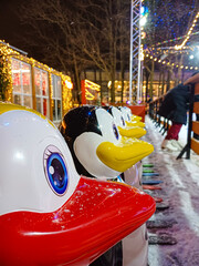 Heads of plastic penguins for children's skating are lined up near an ice rink on a winter evening during a snowfall, illuminated by lights. 