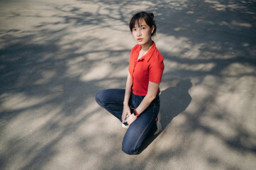 Wall Mural - Asian young woman wearing red shirt and jeans kneels on sunlit pavement, surrounded by tree shadows, with calm and thoughtful expression