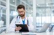 © Liubomir - Concentrated doctor with beard and glasses is sitting at a desk and carefully reviewing patient information on a clipboard in his office