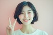 © Маргарита Вайс - pretty cheerful asian young woman with short hair showing peace or victory gesture on pastel pink background