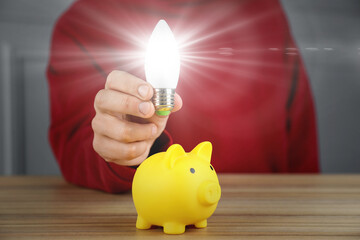 Wall Mural - Man holding glowing light bulb over piggy bank at wooden table, closeup. Energy saving concept