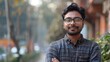 © Prostock-studio - A young man with glasses and a stylish shirt stands confidently, smiling in an urban park. The background features trees and buildings, capturing a peaceful afternoon atmosphere.