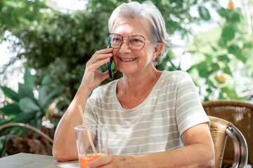 Wall Mural - Happy attractive senior woman sitting in outdoor in a cafeteria table talking at mobile phone, elderly lady relaxing enjoying tech and social