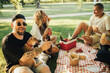 © polinaloves - Happy people having picnic with hot dogs and cold drinks, sitting on a plaid in a park.