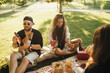 © polinaloves - Happy people having picnic with hot dogs and cold drinks, sitting on a plaid in a park.