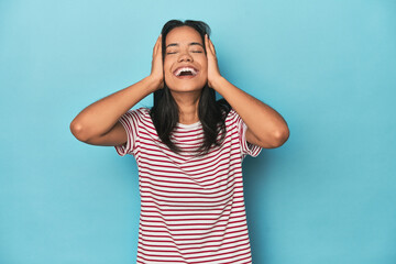 Wall Mural - Filipina young woman on blue studio laughs joyfully keeping hands on head. Happiness concept.