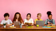 © Prostock-studio - Multiethnic children with electronic devices and books studying at table over pink background, free space. Panorama