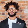 © Vertigo Images - Confident young Black man with a beard and glasses holds a clipboard against a brick wall.
