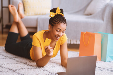 Poster - Online Shopping Concept. Excited african american lady showing credit card and using laptop, ordering clothes and purchases via internet, lying on the floor carpet in living room at home