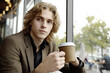 © Athichai - A young professional in casual attire sitting by a large office window, holding a cup of coffee while looking outside, deep in thought. The natural lighting creates a calm and inspiring mood