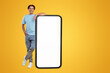 © Prostock-studio - Full body length of smiling casual guy standing and leaning on huge giant cell phone with empty white screen template, orange studio background. Mock up for app website, free space for advertising