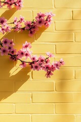 Poster - Delicate pink flowers climb a vibrant yellow brick wall, basking in warm sunshine. Shadows dance softly, creating a serene springtime atmosphere filled with beauty and life