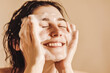 © NARM Applied - Smiling woman enjoying a refreshing face wash, lathered with foam, on neutral background.