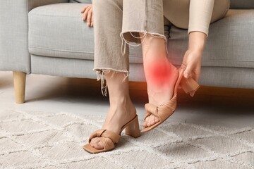 Young woman with pain in ankle taking off uncomfortable shoes at home