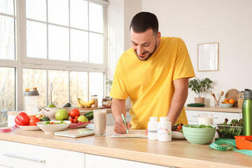 Wall Mural - Handsome young man with fresh vegetables and vitamins making meal plan in kitchen. Healthy food concept