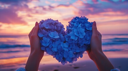 Poster - A woman hands carefully shape blue hydrangea flowers into a heart at sunset on a beach, creating a beautiful contrast with the colorful sky.