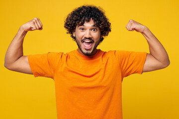 Wall Mural - Young strong sporty fitness happy Indian man he wear orange t-shirt casual clothes showing biceps muscles on hand demonstrating strength power isolated on plain yellow background. Lifestyle concept.