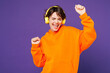 © ViDi Studio - Young smiling fun happy Caucasian woman she wear orange hoody casual clothes listen to music in headphones raise up hands dance isolated on plain purple background studio portrait. Lifestyle concept.
