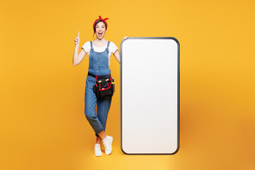 Poster - Full body young employee handyman woman wear overalls belt big blank screen mobile cell phone point finger up isolated on plain yellow background. Instruments for renovation room. Repair home concept.