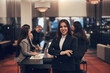 © Flamingo Images - Smiling young businesswoman standing in an office lounge with her arms crossed. Her colleagues are in the background. They held a meeting