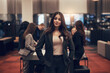 © Flamingo Images - Young businesswoman standing in an office lobby with diverse coworkers in the background. She looks directly into the camera while smiling