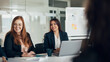 © Flamingo Images - Smiling diverse businesswomen sitting at a conference table during an office meeting. There is a whiteboard in the background with post it notes on it.
