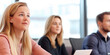 © Vasili - A young woman attentively listening in a business seminar, surrounded by colleagues. The bright and professional setting emphasizes focus, engagement, and corporate learning.